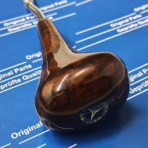 以下が含まれることがあります： 銀色の金属製のインサートとメルセデス・ベンツのロゴが付いた、濃い茶色の木製のシフトノブ。ノブは、白い文字で「Original Parts Geprüfte Qualität」と書かれた青い背景の上にあります。