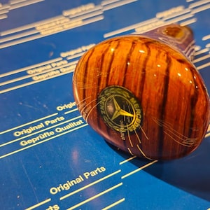 Può includere: Un pomello del cambio in legno con un logo Mercedes-Benz al centro. Il pomello è marrone con una venatura del legno e ha una finitura nera e argentata.