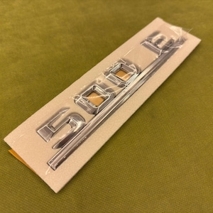 Puede incluir: Emblema "SDE" plateado sobre un respaldo beige, sellado en plástico transparente. El emblema es metálico y tiene una superficie reflectante. El fondo es de color verde oliva liso.