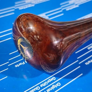 May include: A brown wooden gear shift knob with a gold Mercedes-Benz logo. The knob is on a blue surface.