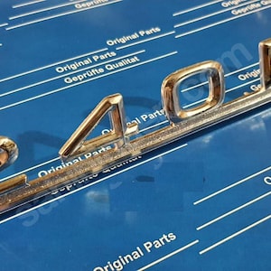 Peut inclure: Un emblème en métal chromé avec le numéro "240D" en relief. L'emblème est conçu pour une voiture et a une forme longue et étroite.