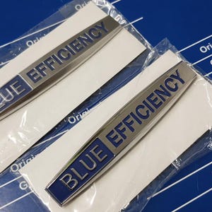 May include: Two silver and blue "BLUE EFFICIENCY" automotive emblems, packaged on white cards, are displayed against a blue background with text. The emblems are rectangular with the text in blue.