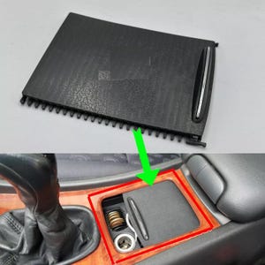 Puede incluir: Un cenicero negro para la consola del coche con una superficie texturizada. El cenicero tiene un acento plateado y se muestra tanto abierto como cerrado. La imagen también muestra el interior del coche, incluyendo la palanca de cambios y la moldura de madera.
