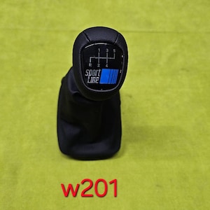 May include: Black car gear shift knob with a blue and white Sport Line design. The shift pattern is visible, with numbers and lines indicating gear positions. The knob is set against a yellow-green background.