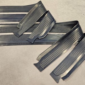 May include: Several dark grey metal trim pieces, some straight and some curved, with a ribbed texture. The pieces are arranged on a light-coloured surface, possibly for automotive or decorative use.
