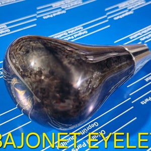 Puede incluir: Una palanca de cambios negra y plateada con un patrón de veta de madera. La palanca tiene una parte superior de metal plateada y una base negra. El texto "BAJONET EYELET" está impreso en la base.
