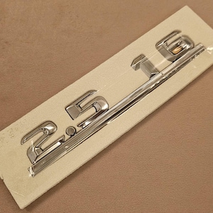 Puede incluir: Un emblema cromado con el número "2.5" se muestra sobre un soporte rectangular blanquecino. Probablemente para un modelo de vehículo. La superficie reflectante del emblema contrasta con el fondo beige liso.