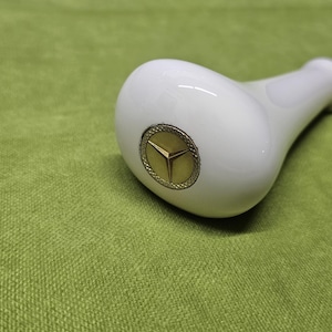 May include: A white, glossy gear shift knob with a gold emblem of a three-pointed star. The knob is set against a green textured background, suggesting a car interior accessory.