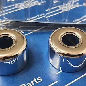 Op de afbeelding: Twee glanzende verchroomde metalen doppen met een rond gat in het midden. De doppen liggen op een blauw oppervlak met witte tekst die "Original Parts" en "Geprüfte Qualität" luidt.