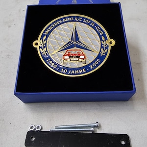 Puede incluir: Un emblema de metal dorado con un logotipo de estrella azul Mercedes-Benz y un coche rojo dentro de un círculo blanco. El emblema tiene el texto "MERCEDES-BENZ R/C 107 SL-CLUB 1990-10 JAHRE-2000" alrededor del exterior.