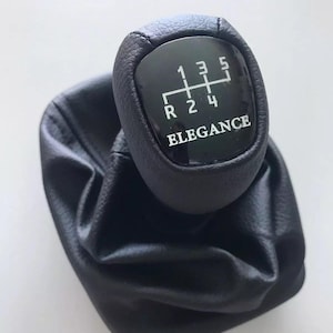 以下が含まれることがあります： 黒い革製のシフトノブで、黒いプラスチック製のトップが付いています。トップには、白い数字の1、2、3、4、5と、リバースを表すRの文字があります。トップには、白い文字で「ELEGANCE」という言葉も書かれています。