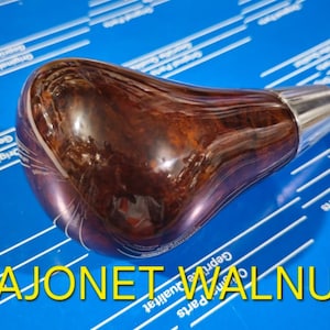 Könnte beinhalten: Ein brauner Holz-Schalthebelknauf mit einem silbernen Metallkopf. Der Knauf hat eine Holzmaserung und ist mit "BAJONET WALNUT" beschriftet.