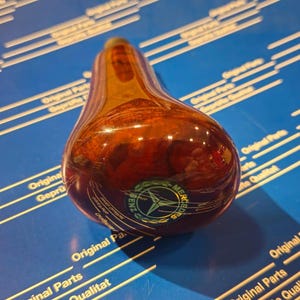 Può includere: Un pomello del cambio in legno marrone con un logo Mercedes-Benz argentato. Il pomello è su una superficie blu con testo bianco.