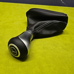 May include: A black leather gear shift knob with a silver base and a circular design. The knob is on a green surface. w211