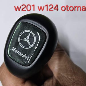 以下が含まれることがあります： 黒いシフトノブに銀色のメルセデス・ベンツのロゴと黒い背景に「メルセデス」の文字。ノブの上に赤い文字で「w201 w124 otomatik」と書かれている。