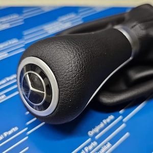 May include: A black leather gear shift knob with a silver trim and a Mercedes-Benz logo. The gear shift is on a blue background with the words "Original Parts" printed on it.