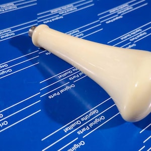 Peut inclure: Un pommeau de levier de vitesse en céramique blanche avec une forme arrondie et bulbeuse. Le pommeau a une finition lisse et brillante.