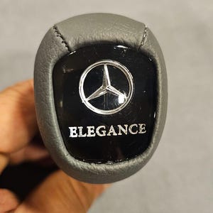 Può includere: Un pomello del cambio in pelle grigia con un centro nero lucido. Il centro presenta il logo Mercedes-Benz e la parola "ELEGANCE" in lettere argentate. Il pomello è progettato per un veicolo.