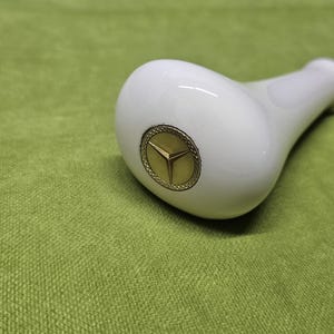 May include: A white, glossy gear shift knob with a gold emblem of a three-pointed star. The knob is set against a green textured background, suggesting a car interior accessory.