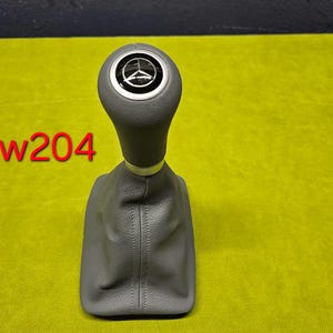 Peut inclure: Pommeau de levier de vitesses en cuir gris avec une garniture argentée et un logo Mercedes-Benz sur le dessus. Le pommeau est étiqueté "w204".