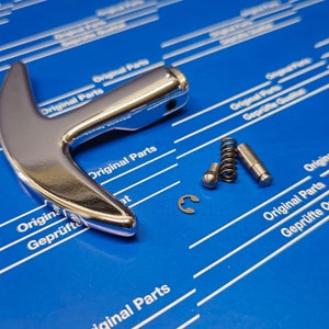 以下が含まれることがあります： T字型のデザインの銀色の金属製のハンドル。ハンドルは、「Original Parts Geprüfte Qualität」という白い文字が書かれた青い背景の上にあります。