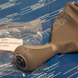 Può includere: Fodera per leva del cambio in pelle grigia con pomello del cambio in plastica grigia. Il pomello presenta il logo Mercedes-Benz e il testo "W211 VITES KORUĞU KOMPLE GRI DERI" è stampato su un'etichetta bianca.