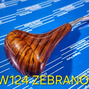 以下が含まれることがあります： 茶色と黒のゼブラウッド模様のウッド製のシフトノブ。ノブは銀色の金属製のシフトレバーに取り付けられています。画像には「W124 ZEBRANO」というテキストが表示されています。