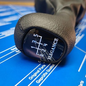 Könnte beinhalten: Schwarzer Leder-Schalthebelknauf mit silberner 'Elegance'-Inschrift und einem schwarz-weißen Schaltmuster. Das Schaltmuster zeigt die Zahlen 1, 2, 3, 4, 5 und R.