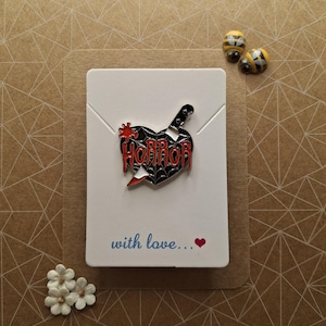 May include: A heart-shaped enamel pin with the word "HORROR" and a knife design. The pin is black and red. It is displayed on a white card with the text "with love..." and a heart. Two bee pins and white flowers are also visible.