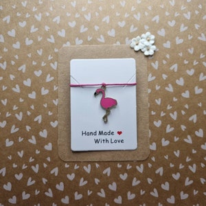 May include: A white card with a pink flamingo charm hanging from a pink string. The card says "Hand Made With Love" and has a small red heart.