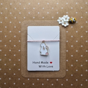 Può includere: Un ciondolo a forma di coniglio bianco con dettagli rosa è appeso a un filo rosa, presentato su una carta bianca. La carta è su uno sfondo marrone a pois bianchi. La carta recita "Hand Made With Love".