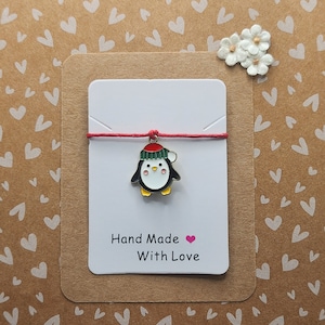 Può includere: Un biglietto bianco con un filo rosso legato intorno. Il biglietto ha un piccolo ciondolo a forma di pinguino con un cappello rosso. Il biglietto dice "Hand Made With Love" con un cuore rosa.