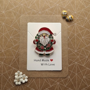 Pode incluir: Um pin com tema natalino com o Pai Natal em fato e chapéu vermelhos, barba branca e uma grinalda verde. O pin está num cartão branco com o texto "Hand Made With Love". Também estão presentes dois pins em forma de abelha e enfeites de flores brancas.