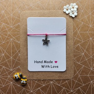 May include: A handmade bracelet with a pink cord and a silver flower charm. The bracelet is displayed on a white card with the text "Hand Made With Love" and a small pink heart. Two small bee-shaped beads are also present.
