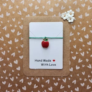Pode incluir: Um cartão branco com um amuleto de maçã vermelha em um cordão verde. O cartão diz "Hand Made with Love" e tem um coração vermelho.