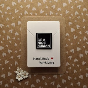 May include: Black and white enamel pin with the text "BE A NICE HUMAN" on a white background. The pin is displayed on a white card with the text "Hand Made with Love" in black and a red heart.