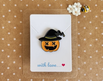 May include: A black and orange enamel pin featuring a pumpkin wearing a witch's hat. The pumpkin has a smiling face with X's for eyes. The pin is on a white card with the text "with love..." in blue.