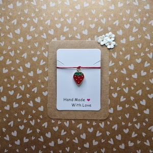 May include: Red and green strawberry charm on a red string bracelet. The bracelet is displayed on a card with the text "Hand Made With Love".