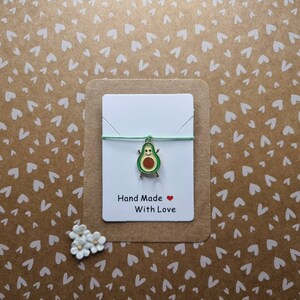 May include: A green avocado charm on a light green string bracelet. The bracelet is displayed on a white card with the text "Hand Made With Love" and a red heart.