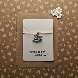 May include: A handmade card with a silver snail charm on a pink string. The card reads "Hand Made With Love" in black text with a red heart. The background is brown with a white heart pattern, and there are small white flowers.