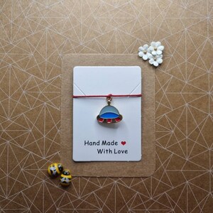 May include: A handmade charm bracelet with a UFO charm in blue, red, and silver. The charm hangs from a red cord, presented on a white card with the text "Hand Made With Love". Two bee-shaped decorative pins are also visible in the image.