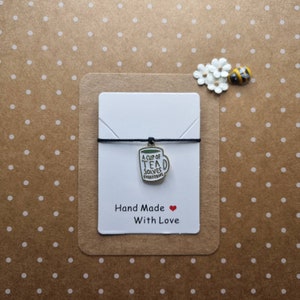 May include: A white card with black text that reads "A cup of tea solves everything" and "Hand Made with Love". A black string with a silver charm of a teacup with the same text is attached to the card.