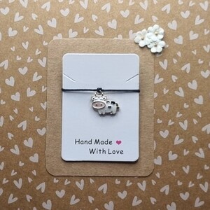May include: A white and black cow charm on a black string bracelet. The bracelet is on a white card with the text "Hand Made With Love".