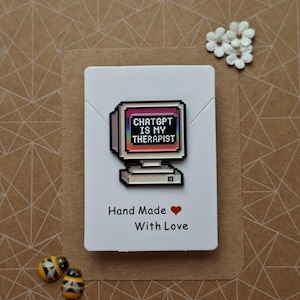 May include: A rectangular enamel pin shaped like a vintage computer monitor with a black screen displaying the pixelated white text CHATGPT IS MY THERAPIST over a rainbow gradient background. The pin is attached to a white card with the text Hand Made.