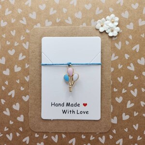 May include: A blue string bracelet with a charm of three balloons in pink, blue, and white. The charm is made of gold metal. The card reads "Hand Made with Love".