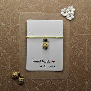 May include: A handmade bracelet with a yellow cord and a small, gold-colored pineapple charm. The bracelet is attached to a white card with the text "Hand Made With Love" and a red heart. Two small bee-shaped beads are also present.