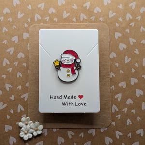 Puede incluir: Un pin de muñeco de nieve con gorro y bufanda rojos, sosteniendo una estrella dorada. El pin está en una tarjeta blanca que dice "Hand Made With Love". El fondo es una tarjeta marrón con formas de corazón blancas y un pequeño grupo de flores blancas.