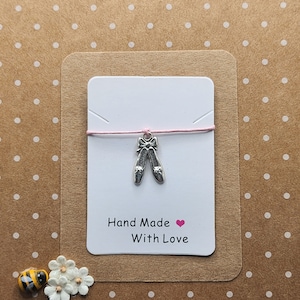 Peut inclure: Une carte blanche avec un cordon rose et un charme de chaussure de ballerine argenté. La carte dit "Hand Made With Love" avec un cœur rose.
