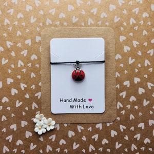 May include: A red ladybug charm on a black string bracelet. The bracelet is displayed on a white card with the text "Hand Made With Love" in black lettering with a pink heart.