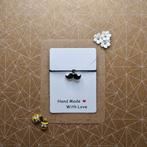 May include: A black string bracelet with a gold charm shaped like a mustache. The bracelet is displayed on a white card with the text "Hand Made With Love" and a red heart.